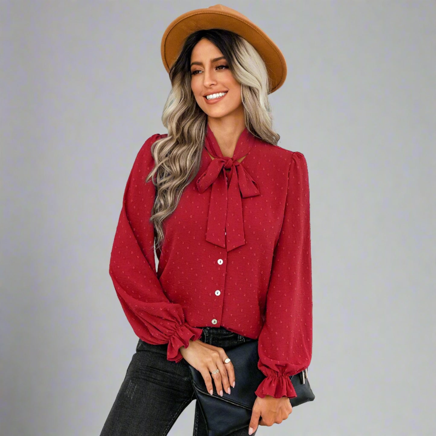 Woman wearing a red bow-tie blouse with a delicate dotted texture, long puffed sleeves, and gathered cuffs, styled with black jeans and a brown hat