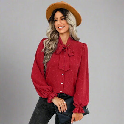 Woman wearing a red bow-tie blouse with a delicate dotted texture, long puffed sleeves, and gathered cuffs, styled with black jeans and a brown hat