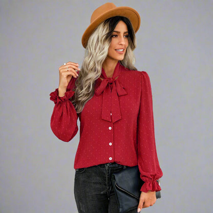 Woman wearing a red bow-tie blouse with a delicate dotted texture, long puffed sleeves, and gathered cuffs, styled with black jeans and a brown hat