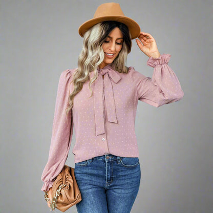 Woman wearing a pink bow-tie blouse with a delicate dotted texture, long puffed sleeves, and gathered cuffs, styled with black jeans and a brown hat