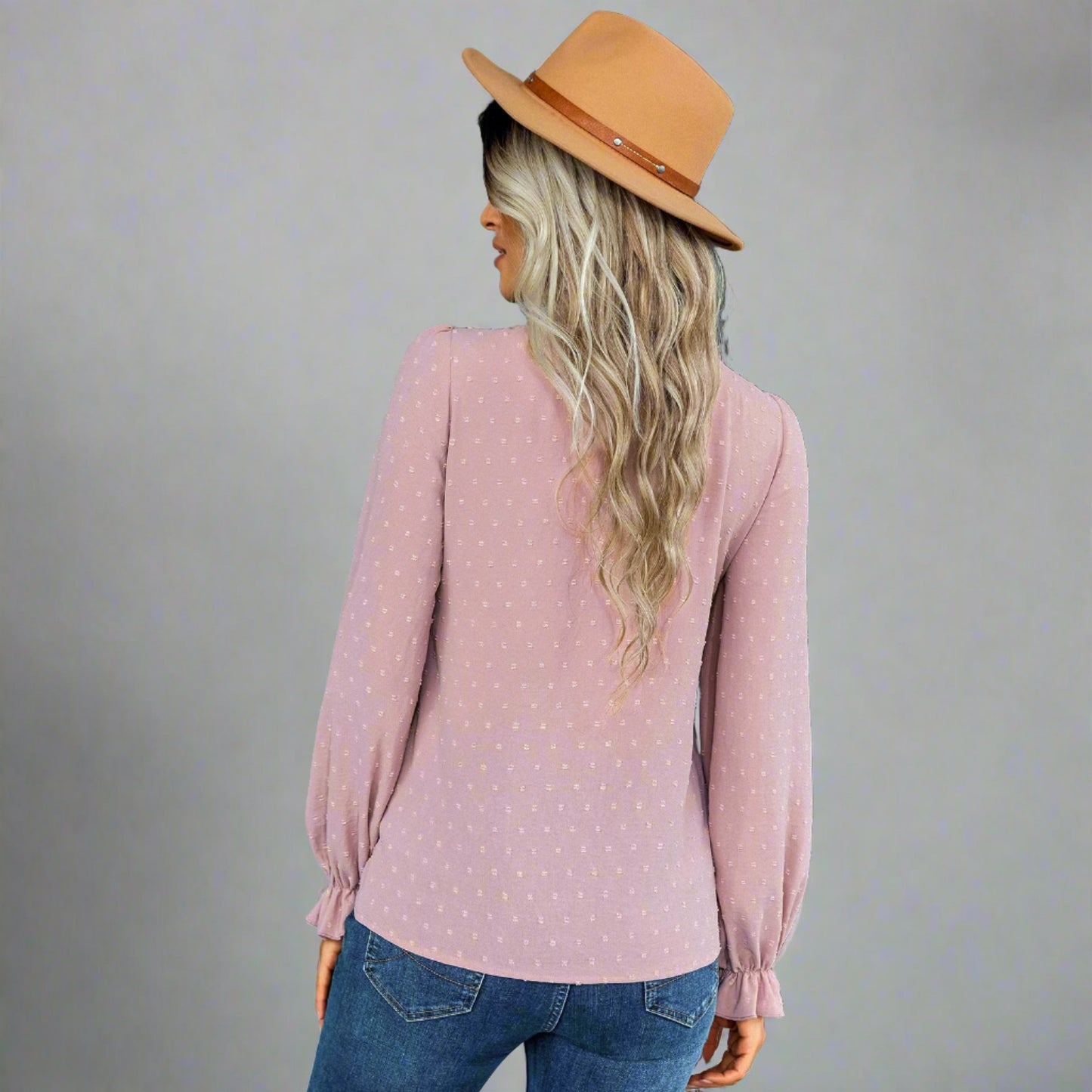 Woman wearing a pink bow-tie blouse with a delicate dotted texture, long puffed sleeves, and gathered cuffs, styled with black jeans and a brown hat