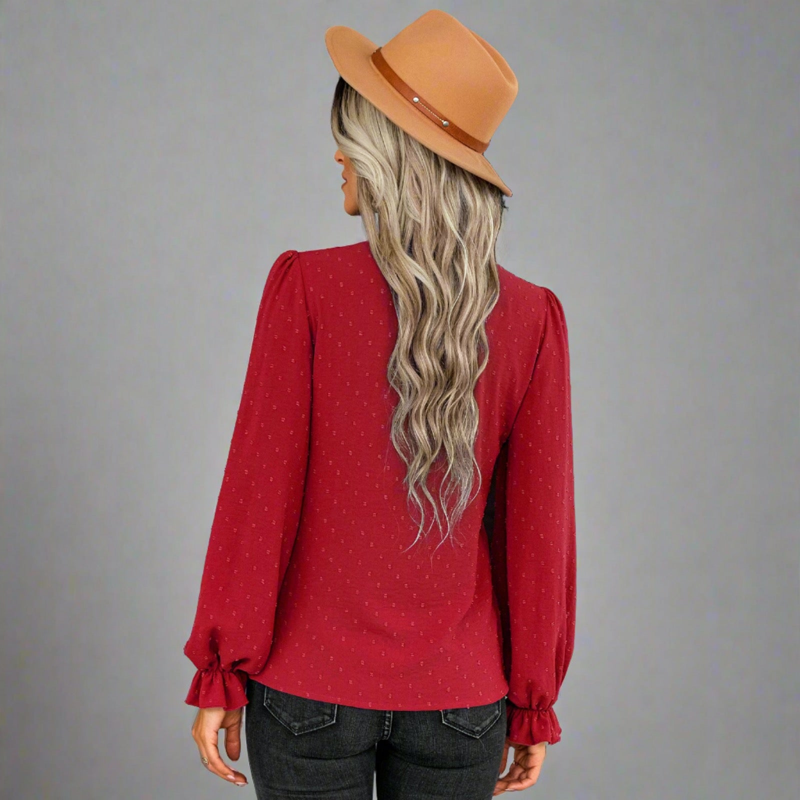 Woman wearing a red bow-tie blouse with a delicate dotted texture, long puffed sleeves, and gathered cuffs, styled with black jeans and a brown hat