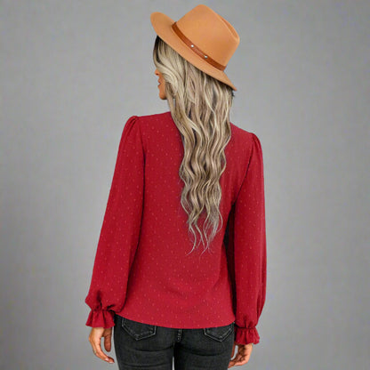 Woman wearing a red bow-tie blouse with a delicate dotted texture, long puffed sleeves, and gathered cuffs, styled with black jeans and a brown hat