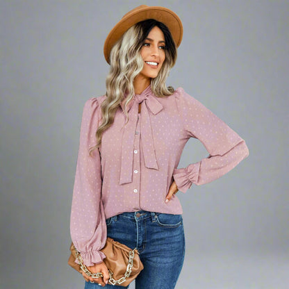 Woman wearing a pink bow-tie blouse with a delicate dotted texture, long puffed sleeves, and gathered cuffs, styled with black jeans and a brown hat