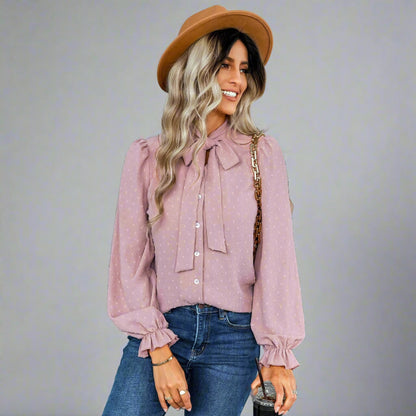 Woman wearing a pink bow-tie blouse with a delicate dotted texture, long puffed sleeves, and gathered cuffs, styled with black jeans and a brown hat